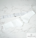 A pristine white Marcella cotton cummerbund with front button detailing and an adjustable back slider strap with metal hooks, displayed on a clean marble table.
