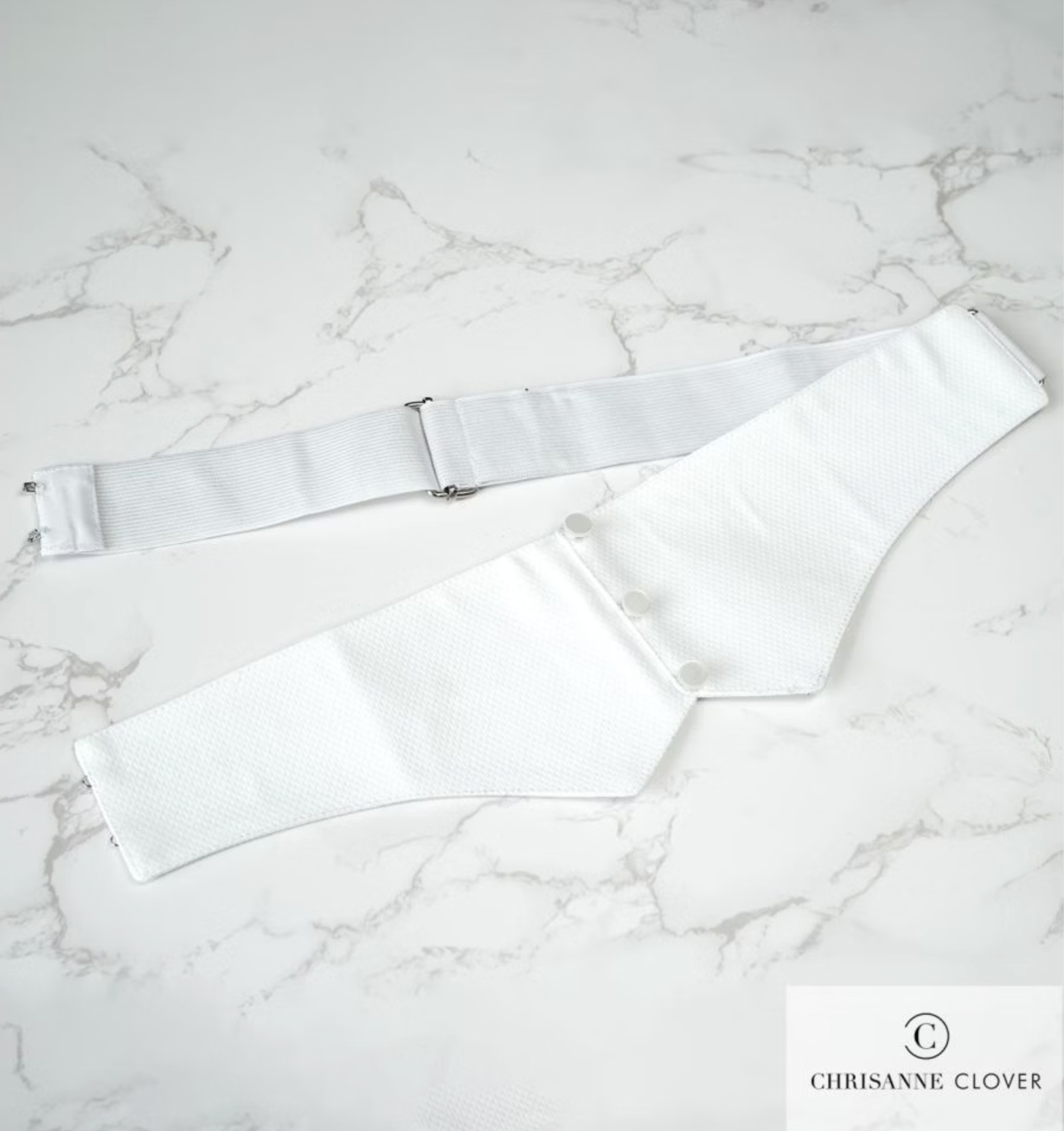 A pristine white Marcella cotton cummerbund with front button detailing and an adjustable back slider strap with metal hooks, displayed on a clean marble table.